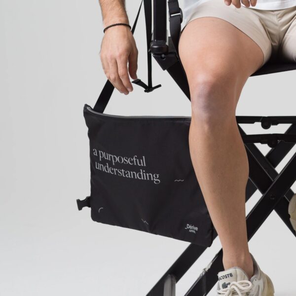 Black Dérive Purposeful Musette held by its strap, showing the side buckle and printed script